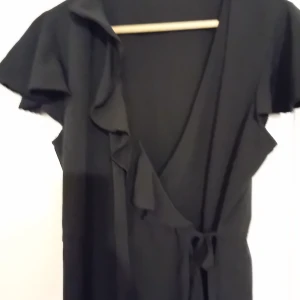 Svart omlott klänning Pernilla Wahlgren  - Svart omlottklänning strl M från Pernilla Wahlgren collection från gekås. Kantad med volang, tunn chiffong liknande material.