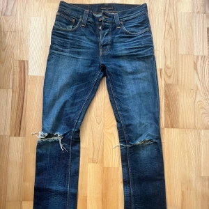 Nudie jeans Grim Tim W31 L32 - Nudie jeans i modell Grim Tim som är slim fit, mid waist, straight leg. Sitter skitsnyggt på men är nu försmå för mig som är 189 cm och 75 kg. Snygga hål över båda knäna, gillar man inte detta går det att lämna in för gratis lagning på Nudies butiker