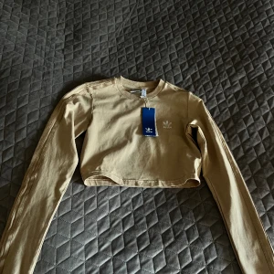 Adidas crop top  - Långärmad top från Adidas Originals Mudd i halsring Broderad logga fram Croppad modell Klassiska tre Adidas streck längs ärmar