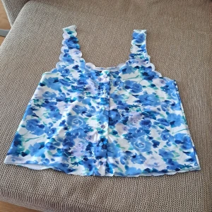 Blommig linnetopp - En blommig linnetopp med blå och vita nyanser. Toppen har breda axelband och knappar framtill. Den har en vågig kantdesign längs axelbanden och halsringningen.