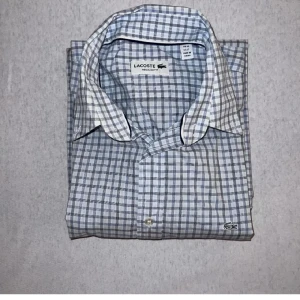 Rutig Skjorta Lacoste - Blårutig skjorta från Lacoste i använt skick. Finns en fläck på vänsterarmen men annars bra skick, fler bilder skickas på förfrågan! Frågor och funderingar är varmt välkomna👍 LIKNANDE PLAGG PÅ PROFILEN💯