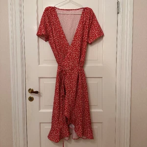 Röd klänning med vita blommor - Röd omlott-klänning med vita blommor. Knyts i midjan med tillhörande band. Går nedanför knäna. Volang längst ned. Står 2XL, men är som en L. Aldrig använd. 