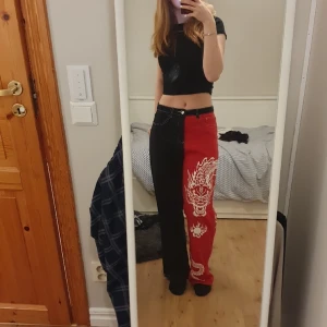 Delade svartröda jeans med drake - Jeans med ett rött ben och ett svartben med en vit drakdetalj på ena benet. Egentligen highwaist men sitter mer som midwaist på mig som har 36. Använda och har tyvärr slitage längst ner på röda benet (bild 3). Annars bra skick. Säljer pga ej min stil.