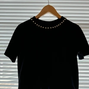 Svart t-shirt med pärlor  - Jag säljer nu denna svarta t-shirt som har vita pärlor framtill vid halsen. (Pärlorna går ej hela vägen runt utan bara framtill) - tröjan är från Zara och är i väldigt gott skick då den knappt är använd 