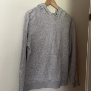 Grå hoodie från H&M - En bekväm grå hoodie från H&M Basic-kollektionen. Tröjan har en huva och en framficka. Den är långärmad och har ribbstickade muddar vid ärmslut och nederkant.