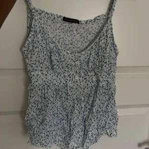 Blommig linnetopp från Brandy Melville - Säljer en söt blommig linnetopp från Brandy Melville. Toppen är vit med små blå blommor och har en v-ringad hals. Den är ärmlös och har knappar framtill. Perfekt för sommaren! superfint skick behöver bara strykas, den har även tunna band på baksidan så man kan knyta den❤️
