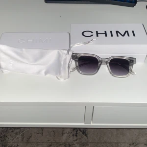 Gråa solglasögon från CHIMI - Säljer ett par snygga gråa solglasögon från CHIMI. De har en rektangulär form och kommer med ett vitt fodral och en vit putsduk. Perfekta för att skydda ögonen mot solen och samtidigt se stiliga ut. Passar både till vardags och fest!