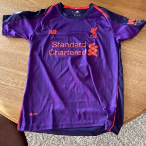 Lila fotbollströja från New Balance - Säljer en snygg lila fotbollströja från liverpool med orange detaljer och LFC-logga på bröstet. Tröjan har korta ärmar och är i mycket bra skick. Perfekt för fotbollsfans eller för att träna i. Materialet är lätt och andas bra.