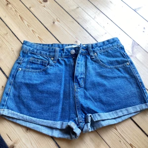 Blå jeansshorts - Säljer ett par snygga blå jeansshorts i storlek M. De är i nyskick och perfekta för sommaren! Shortsen har en normal passform med uppvikta ben och klassisk femficksdesign. De är tillverkade av slitstarkt denim och har en knapp och dragkedja framtill. Perfekta för både vardag och fest!