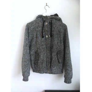 Jacka i hoodie modell, S 36/38, ull, grå, kort, varmfodrad, vinter, höst, alternative, emo - En mörkgrå jacka i ullblandning. Hoodiemodell som stängs med dragkedja och tryckknappar. Två sidfickor och stor huva med dragsko. Finns noppor. Mått i cm: bröstvidd 51; axelbredd 40; längd fram från axeln 58; ärmlängd 60.