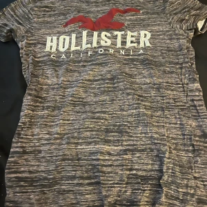 Gråmelerad t-shirt från Hollister - En gråmelerad t-shirt från Hollister med texten 'Hollister California' tryckt på framsidan. T-shirten har korta ärmar och är tillverkad i ett mjukt och bekvämt material.