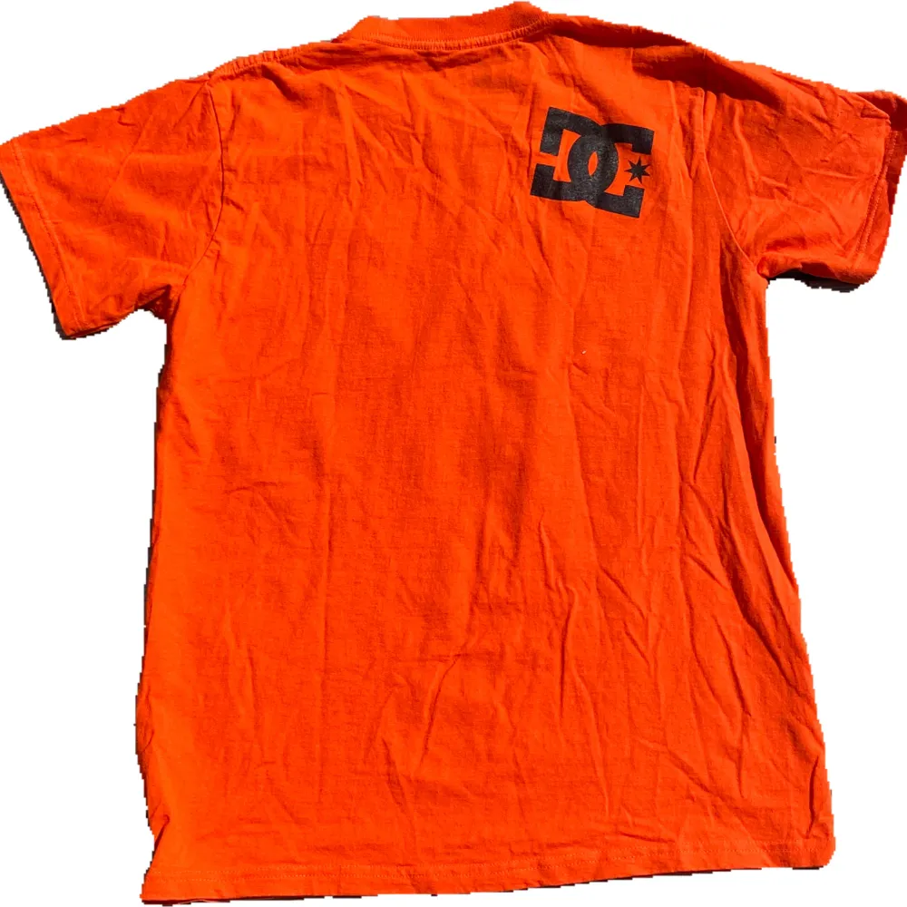 En orange t-shirt från DC med ett stort svart och grått tryck på framsidan och ett mindre svart tryck på baksidan. T-shirten har korta ärmar och en rund halsringning. storlek M sitter som S. skriv för fler frågor! 🐡🐰🐹🐭. T-shirts.