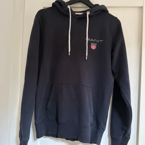 Hoodie GANT, S - Marinblå hoodie från GANT Storlek S Utseende enligt bild Ytterst litet märke på fickan, vilket syns på sista bilden. Stör ej funktionen och jag själv har inte sett det förrän jag granskade den nu. 