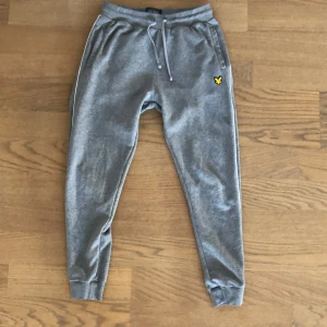 Grå mjukisbyxor från Lyle & Scott - Ett par grå mjukisbyxor från Lyle & Scott med resårmidja och dragsko. Byxorna har två sidofickor och en broderad gul logotyp på vänster ben. Byxorna är i bra skick och lite använda! 