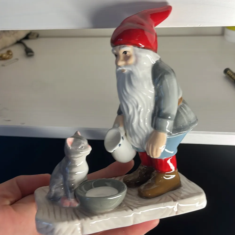 En detaljerad keramikfigur föreställande en tomte med röd luva, grå jacka, blå byxor och bruna stövlar. Tomten håller en vit kanna och står bredvid en grå katt som sitter vid en skål. Figuren är målad i naturliga färger och har en glansig yta.. Accessoarer.