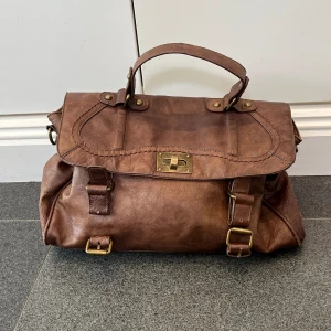 Brun handväska  - Brun handväska från Ulrika. Väl använd men ganska fint skick, kom privat för fler bilder. Ca 35 cm bred och ca 19 cm hög. Köpt secondhand och hon jag köpte den av sa att det var äkta skinn.