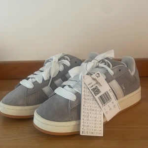 Helt nya gråa adidas campus 38 - Helt nya Adidas campus i strl 38. Detta är dam modellen vilket gör att man får de super fina breda skosnörena till.   Aldrig använda, nya med prislapp. Kartong finns den är däremot skadad från frakten. Orderbekräftelse finns även om man vill se bild.