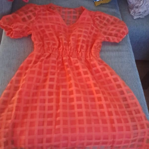 Orange kortärmad klänning med rutmönster - En orange kortärmad klänning med ett genomskinligt rutmönster. Klänningen har en v-ringad hals och en elastisk midja som ger en bekväm passform. Den är kort i längden och har puffärmar. oanvänd!