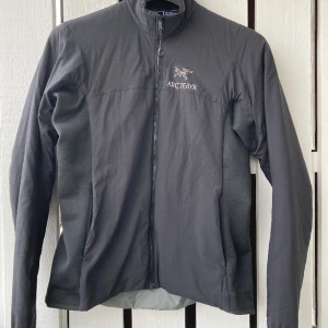 Arc'teryx Bust Buste mellanlagerjacka - Arc'teryx mellanlagerjacka, storlek M, svart. Perfekt under en skaljacka eller som den är. Tunnt isolerad, stretchiga partier i sidorna med fleece på insidan. Fleeceklädd krage. Två fickor med dragkedja framtill och en innerficka med dragkedja.