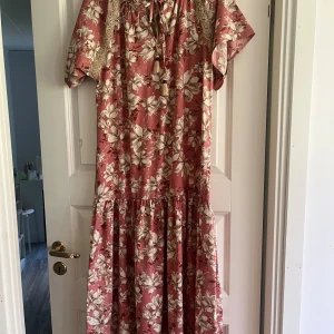 Blommig klänning från Meisie - En vacker blommig klänning från Meisie i rosa och beiga toner. Klänningen har korta ärmar och en lös passform med volangdetaljer. Den har även spetsdetaljer och en knytning vid halsen.