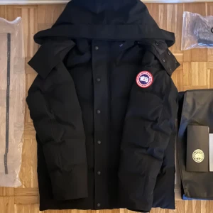 Svart dunjacka från Canada Goose - En svart dunjacka från Canada Goose med en hög krage och huva. Jackan har en broderad logotyp på vänster ärm och stängs med både dragkedja och knappar. Insidan är quiltad för extra värme. storlek M. har en liten defekt på höger sidan av luvan se sista bilden.