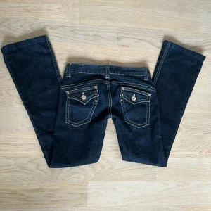 Lågmidjade jeans - Mörkblåa/svarta jeans med vita sömmar. Snygga bakfickor! Innebenslängd: 77cm. Midjemått tvärs över: 38 cm. Byxorna är uppsydda någon cm. De är lite slitna vid hälarna men det märks inte vid användning.