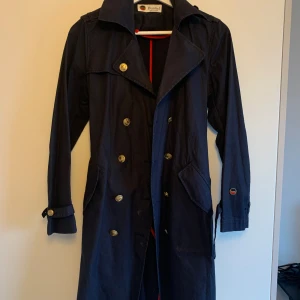 Trenchcoat från Busnel  - Marinblå trenchcoat med röda detaljer från Busnel. Bra använt skick. En knapp saknas vid vänster ärm (bild 3) men kan enkelt sys dit. Perfekt vårjacka. 