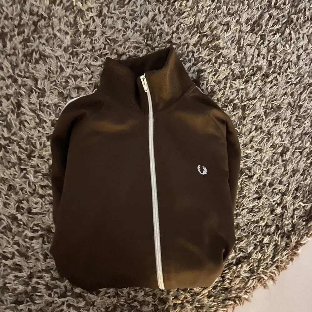 Detta är en brun track jacket med vita detaljer. Den har en dragkedja framtill, vita ränder längs ärmarna och ett litet Fred Perry-märke på bröstet. Designen är stilren och sportig.. Neuletakit & Villapaidat.