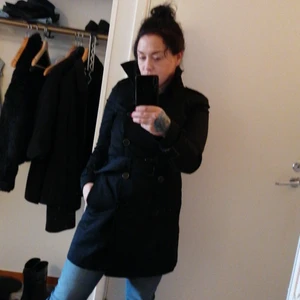 Trenchcoat halvlång  - Svart, stilren, dubbelknäppt trenchcoat med uppvikbar krage och avtagbart skärp (ta av före tvätt!). Bekväm, passar bättre för höst än vinter. Den är figurnära i passformen och S är därför en korrekt beskrivning av storlek, och du får inte plats med någon tjockare tröja under om du inte väljer att bära den öppen eller har en  ganska petit kroppstyp. Den är verkligen snygg, och använd så få gånger att den är i totalt nyskick. I. På mig som är 160 cm är den knälång. Liten puffärm,liten slits bak. 