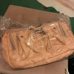 Nunoo rosa väska  - Nunoo rosa/aprikos väska som är knappt använd. Använt den kanske 2 ggr. Har plast osv kvar. Original pris på väskan var 1800kr. Mitt pris är 500kr. Finns ett handtag som man kan sätta på som är längre. Väldigt fint skick! Kan mötas i Stockholm och frakta!