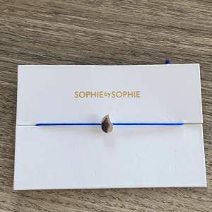 Armband från Sophie By Sophie. WATERAID Bracelet  * Rhodiumpläterat silver med justerbar nylonsnöre. * Längd: 22 cm * Symbol: H. 0,9 cm B. 0,5 cm Färg : blå   