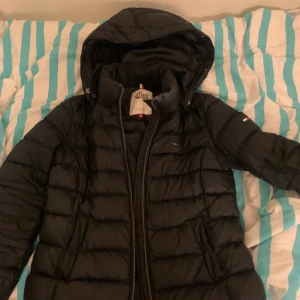Tommy hilfiger vinterjacka dun - Svart Vinterjacka från Tommy hilfiger. Storlek xs. Jackan är i dun. Använd men bra skick, däremot är det en liten trasig bit (vet inte vad jag ska kalla det men kolla på andra och tredje bilden) över vänstra fickan samt vid vänstra armen (detta pga en pin). Dessa syns dock inte alls på långt håll och har inte orsakat några problem för mig.  Nypris 2200 kr. Finns även fluff till jackans luva. Skriv för mer bilder 