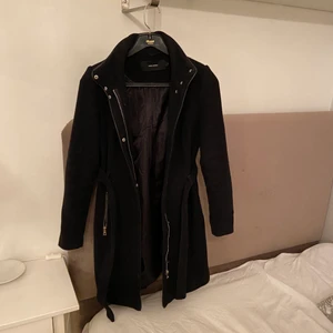 VERO MODA kappa strl S svart - Kappa från VERO MODA i svart. Foder insida tyg utsida. Dragkedja fram och knappar. Skärp i midjan, två fickor fram med dragkedja. Inga slitage. (Frakt exklusive satt pris)