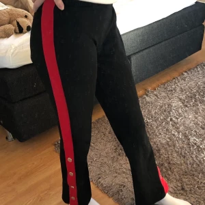 Svarta byxor med röd rand - Ett par jättefina svarta byxor med en röd rand på var sin sida. Knappar längst ner på båda sidorna som går att öppna upp. Säljer då jag aldrig använder de. Byxorna är i jättefint skick, som nya.