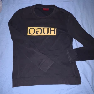 Svart HUGO sweatshirt med gult tryck - Svart sweatshirt från HUGO med lång ärm och rund hals. Inga fläckar och inga hål , det ser ut som en fläck men det kom precis damm på kameran :)