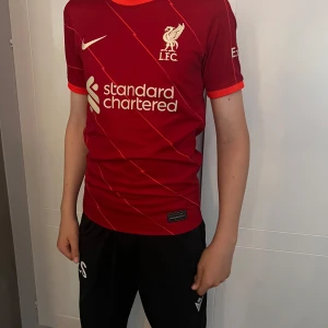 Liverpool FC röd matchtröja Nike - Liverpool FCs officiella matchtröja från Nike i rött med tunna diagonala blixt-ränder. Broderad LFC-logga och Nike swoosh på bröstet, samt sponsortryck framtill. Tröjan är kortärmad och gjord i ett lätt, ventilerande funktionsmaterial.