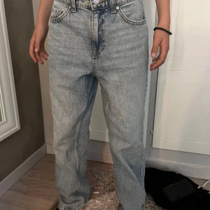 Ljusblå raka jeans med hög midja - Säljer ett par ljusblå jeans med klassisk femficksmodell och raka ben. Jeansen har hög midja och är tillverkade i denim av bomull. Perfekta för en avslappnad och trendig look. Passformen är loose och de har bälteshällor och knappgylf.