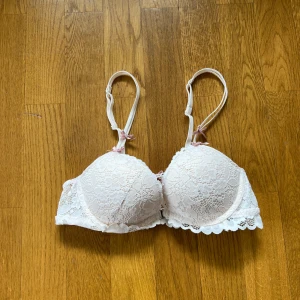 Vit spetsig bh från 6IXTY8IGHT - Supersöt vit bh från 6IXTY8IGHT med spets över hela kuporna och fina små rosetter i ljusrosa. Baksidan har snygg snörning och spetsdetaljer längs kanten. Justerbara axelband och vadderade kupor för extra komfort. Storlek 70A.