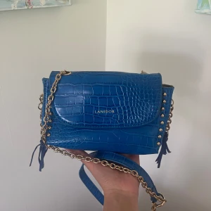 Blå axelväska från Lanidor med kedja - Snygg blå axelväska från Lanidor i croco-mönstrat skinnimitation och dekorativa tofsar. Stängd med ett magnetlås, och insidan har ett fack med dragkedja. Väskan är helt ny men utan prislapp.