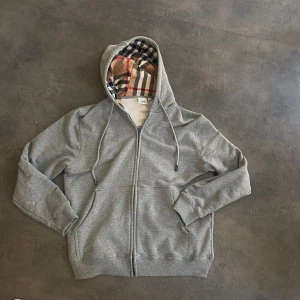 Grå Burberry-Zip - Snygg grå hoodie med dragkedja och snörning i huvan. Insidan av huvan har ett klassiskt rutigt mönster i beige, svart och rött. Tröjan har långa ärmar, ribbade muddar och två fickor framtill. Perfekt för chill dagar eller när du vill vara extra bekväm. 