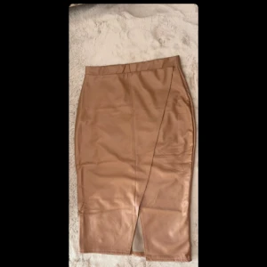 Beige skinnimitation kjol med slits - Snygg beige långkjol i skinnimitation med omlottkänsla och slits framtill. Kjolen har en figurnära passform och stilren design som ger en cool vibe. Perfekt att styla med boots eller sneakers för en trendig look.