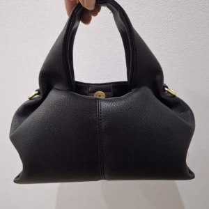 Svart handväska i skinn med axelrem - Säljer en stilren svart handväska i mjukt skinn med både handtag och justerbar axelrem. Väskan har guldfärgade metalldetaljer och en enkel, modern design som passar till många outfits. Perfekt storlek för det viktigaste du behöver ha med dig. Påminner polene väskan!