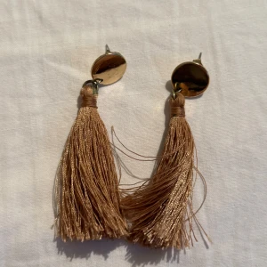 Guldiga örhängen med tofsar - Snygga örhängen med rund, blank guldfärgad platta och långa beige tofsar i glansigt material. Perfekta för att ge en trendig touch till din look. Lätta att bära och riktigt statement.