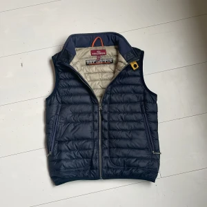 Mörkblå dunväst från Parajumpers - Snygg mörkblå dunväst från Parajumpers med quiltad design och hög krage. Västen har dragkedja framtill, två sidofickor med dragkedja och ikonisk PJS-logga på ryggen. Insidan är ljusbeige och västen har en orange hängare i nacken. Perfekt för lager-på-lager-stil. Kedjan på en ficka har gått av på ena zippen men går att fixa om man är bra på att peta. Märket där bak på ryggen är lite lös men syns knappt och man kan sy lite. Om vi vill ha bild på nån av de är det bara att skriva till mig.