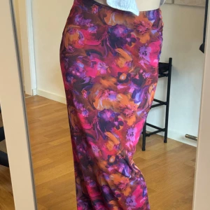 Färgstark långkjol från PrettyLittleThing - Säljer en långkjol från PrettyLittleThing med färgstarkt blommigt mönster i lila, rosa och orange toner. Kjolen har en figurnära passform och en liten slits bak. Materialet känns lätt och följsamt, perfekt för dig som vill sticka ut med en cool look.