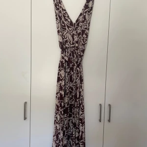 Brun beige långklänning med blommigt mönster från Lindex - Säljer en brun långklänning från Lindex med beige blommigt mönster. Klänningen har v-ringning, är ärmlös och har ett matchande knytband i midjan. Perfekt för dig som gillar flowiga och snygga plagg med fina detaljer.