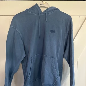Blå hoodie från Levi's med huva - Säljer en blå hoodie från Levi's med broderad logga på bröstet. Tröjan har huva med snörning och en stor känguruficka framtill. Perfekt för dig som gillar en avslappnad och stilren look. Långärmad och i mjuk bomull.