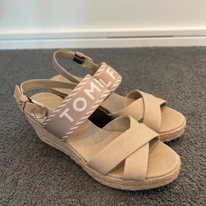 Beige sandaler med kilklack Tommy Hilfiger - Snygga beige klacksf från Tommy Hilfiger med breda justerbara remmar med guldfärgat spänne och tydlig logga framtill. Skorna har en hög kilklack i rep och öppen tå, perfekta för sommarens alla outfits. Endast använda 1 gång!