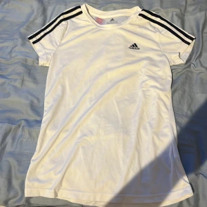 Vit sportig T-shirt från Adidas - Sportig vit T-shirt från Adidas med svarta ränder på axlarna och klassisk logga på bröstet. Tillverkad i lätt och funktionellt material som andas. Kortärmad och med en enkel, rak passform som ger en clean look. Bra för träning. Storlek 170. Passar barn och ungdomar i storlek S,XS. 