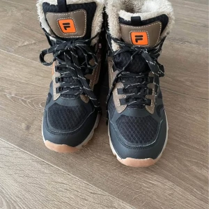 Svarta vinterboots från Fila, stl 40 - Snygga svarta och bruna vinterboots från Fila med grov sula och mysigt foder i beige pälsimitation. Skorna har snörning framtill och detaljer i syntet och mesh för extra komfort. Perfekta för kalla dagar när du vill hålla stilen och värmen.